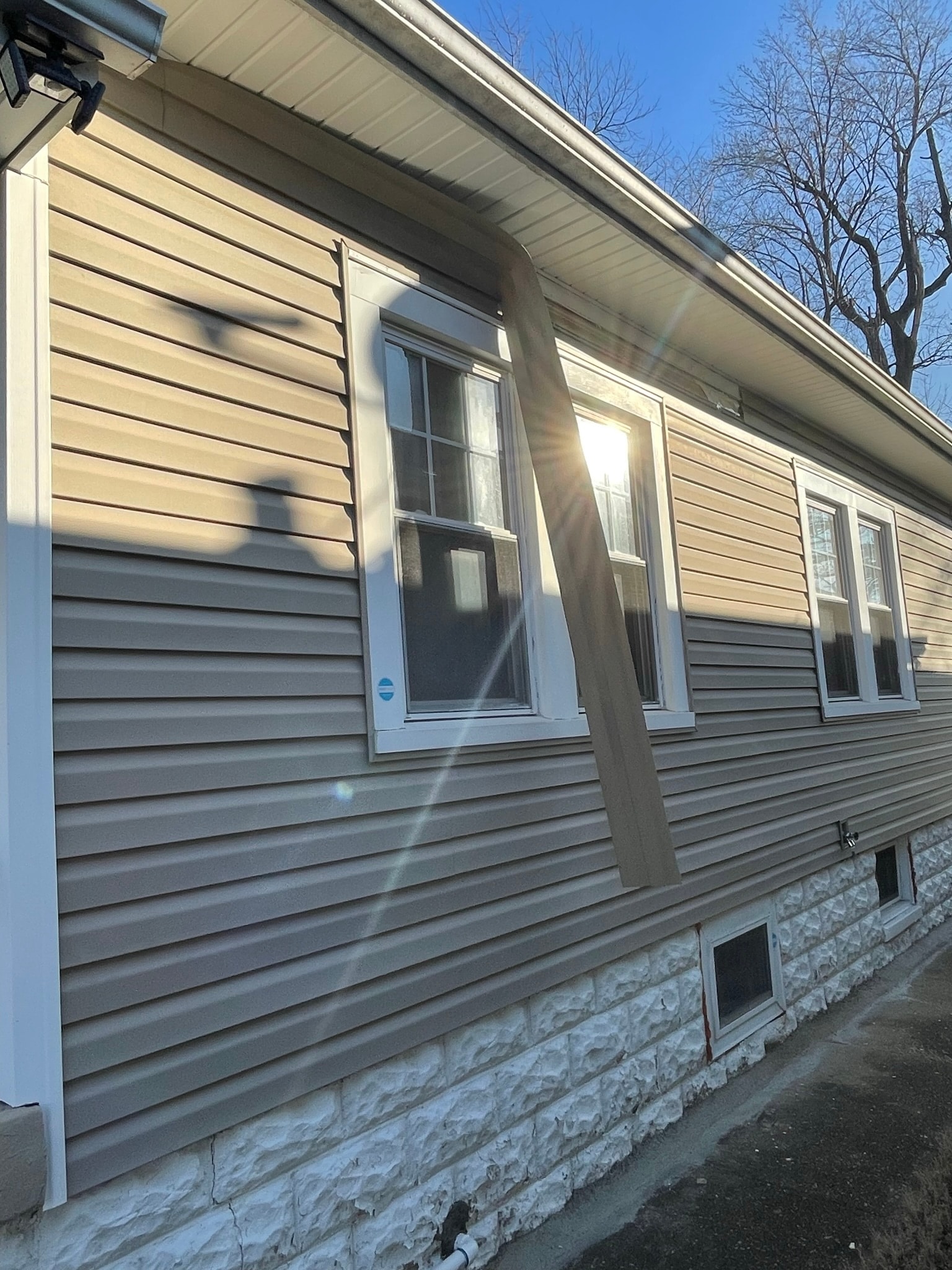 House exterior before repair — damaged corner trim and downspout hanging loose away from tan vinyl siding