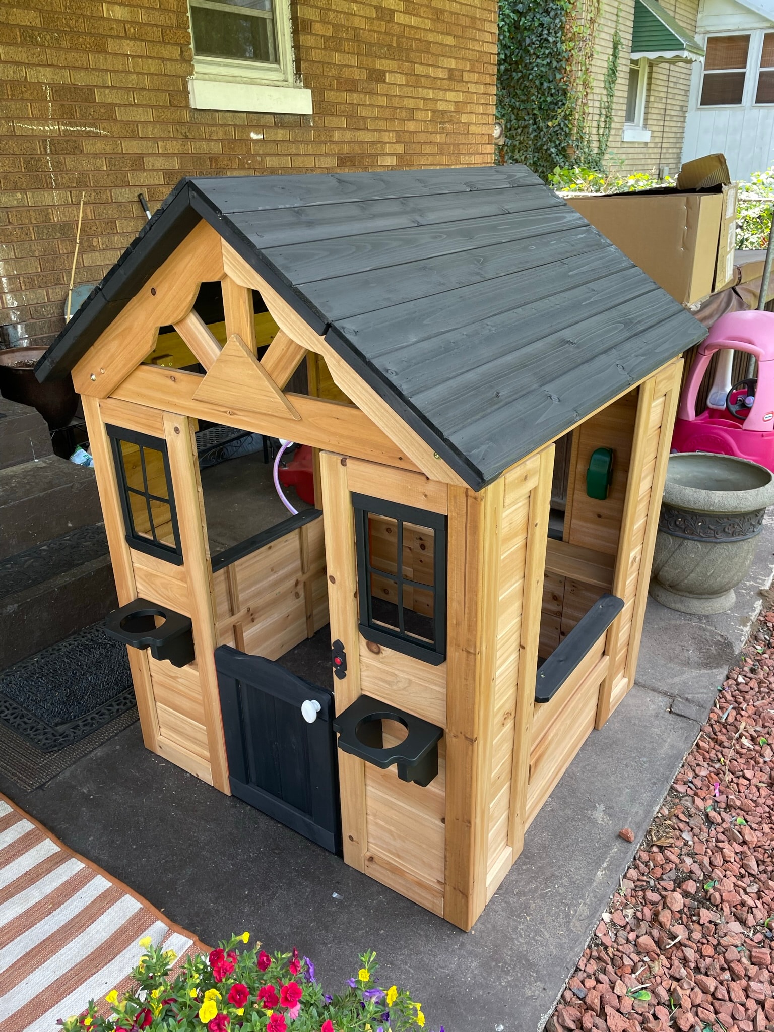 Fully assembled cedar playhouse with dark charcoal roof, black trim door and windows, and decorative gable detail on a backyard patio
