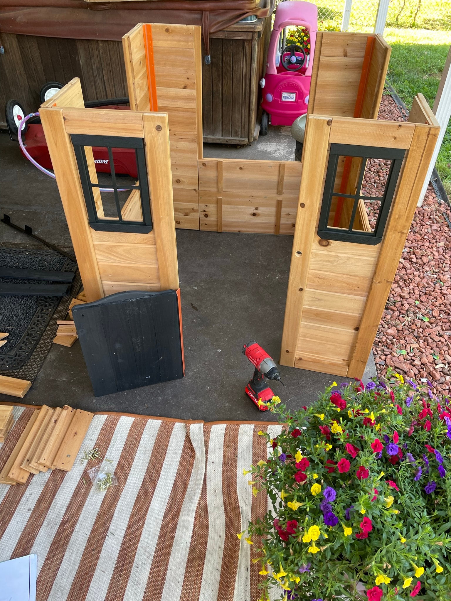 Cedar playhouse mid-assembly — walls and window frames standing without a roof, drill and loose boards on the patio
