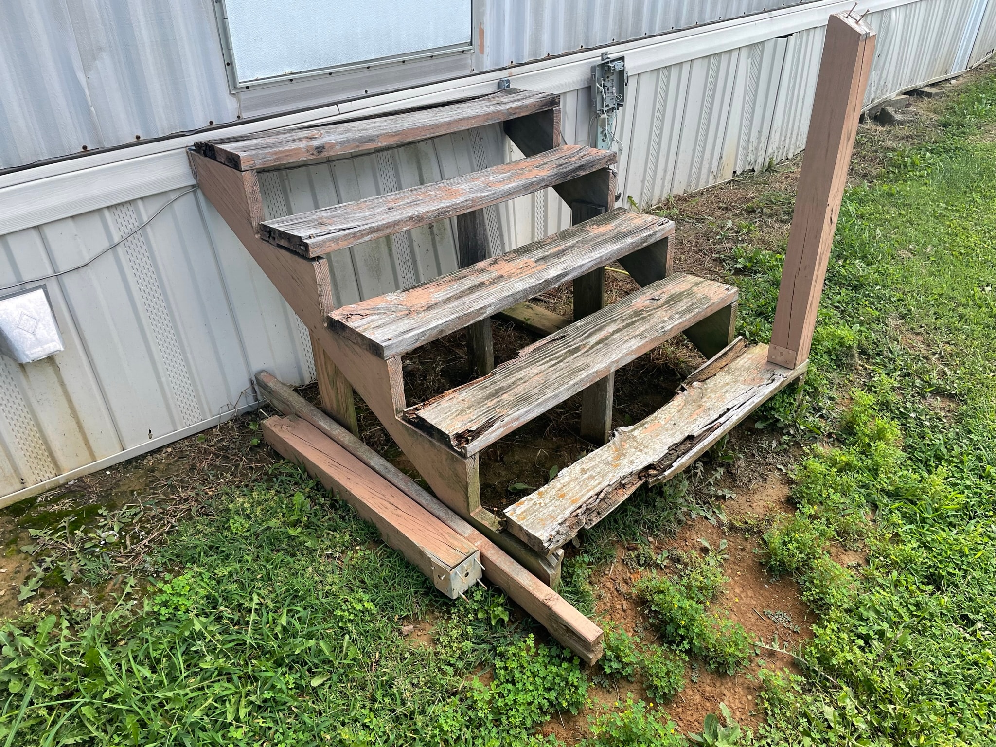 Mobile home exterior before steps replacement — severely rotted and collapsed wooden entry steps with peeling paint alongside metal siding