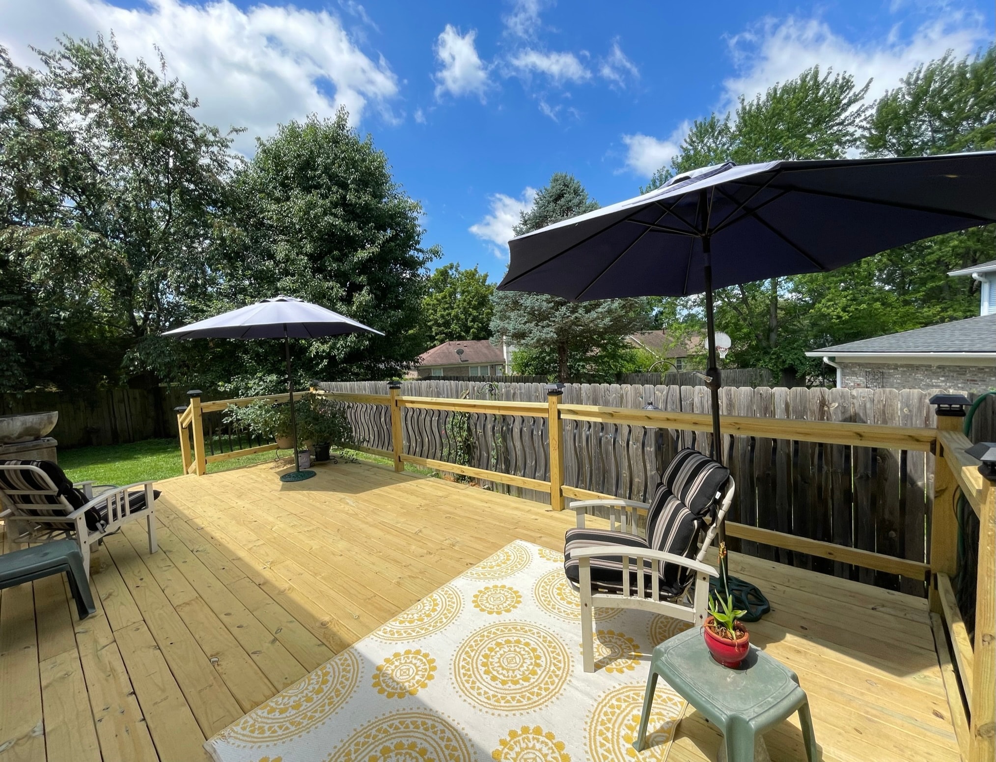 Backyard after deck build — large finished pressure-treated wood deck with decorative iron spindle railings, solar post caps, patio furniture, and navy umbrellas under a sunny blue sky