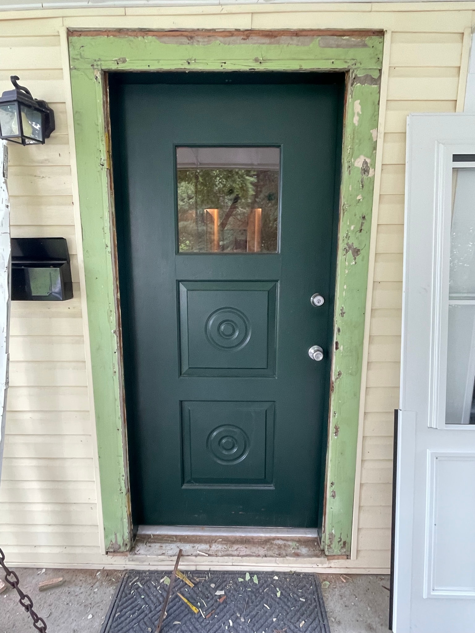Residential front entrance before door replacement — dark green paneled door with peeling green trim frame mid-installation, debris on doorstep