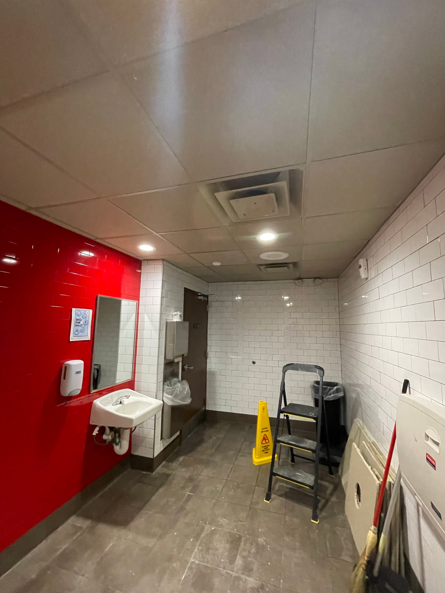 Commercial bathroom after ceiling repair — fully replaced clean flat drop ceiling tiles with proper lighting, step ladder and wet floor sign still on site