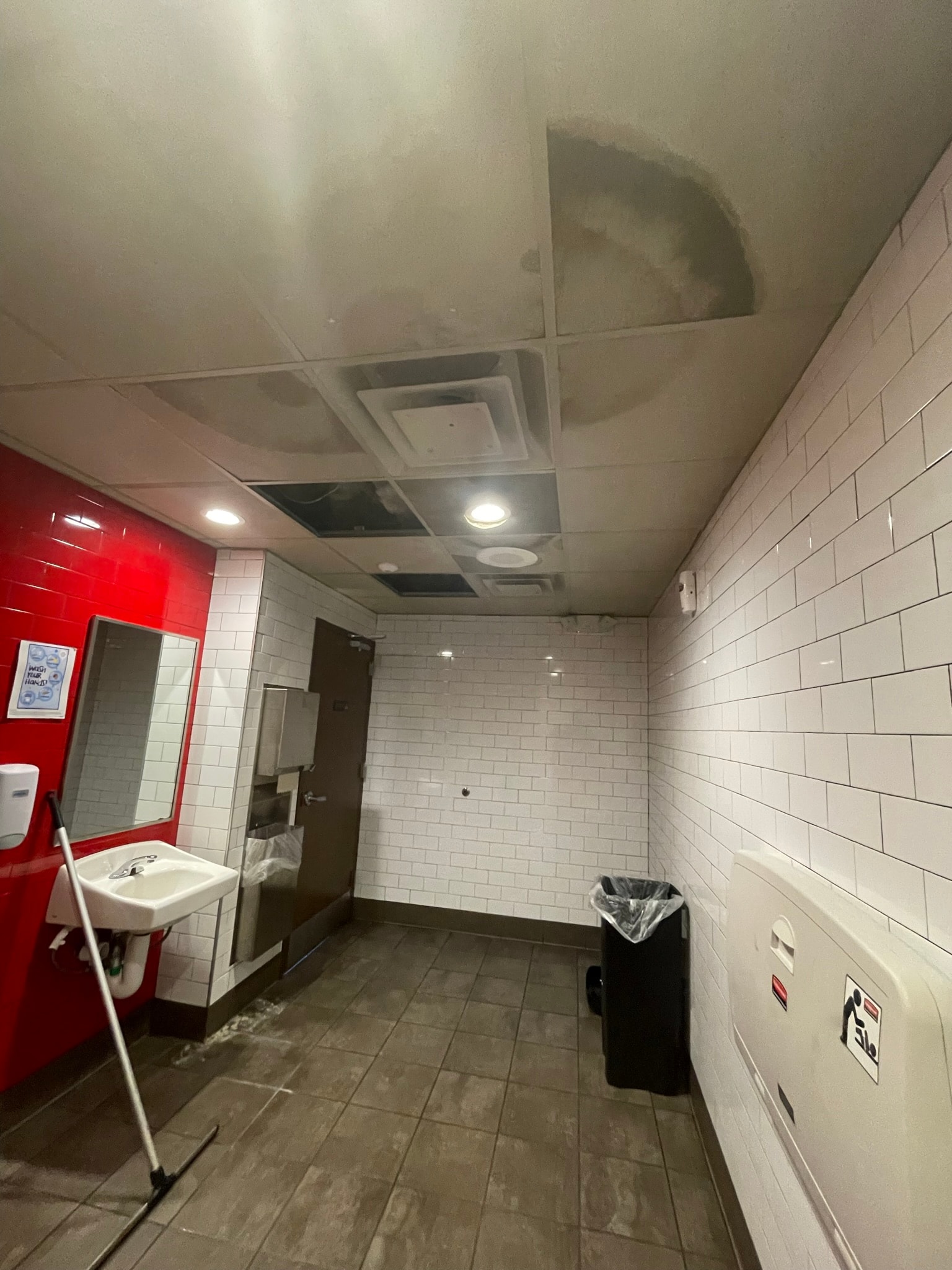 Commercial bathroom before ceiling repair — severely water-damaged sagging drop ceiling tiles with dark staining and mop on wet floor
