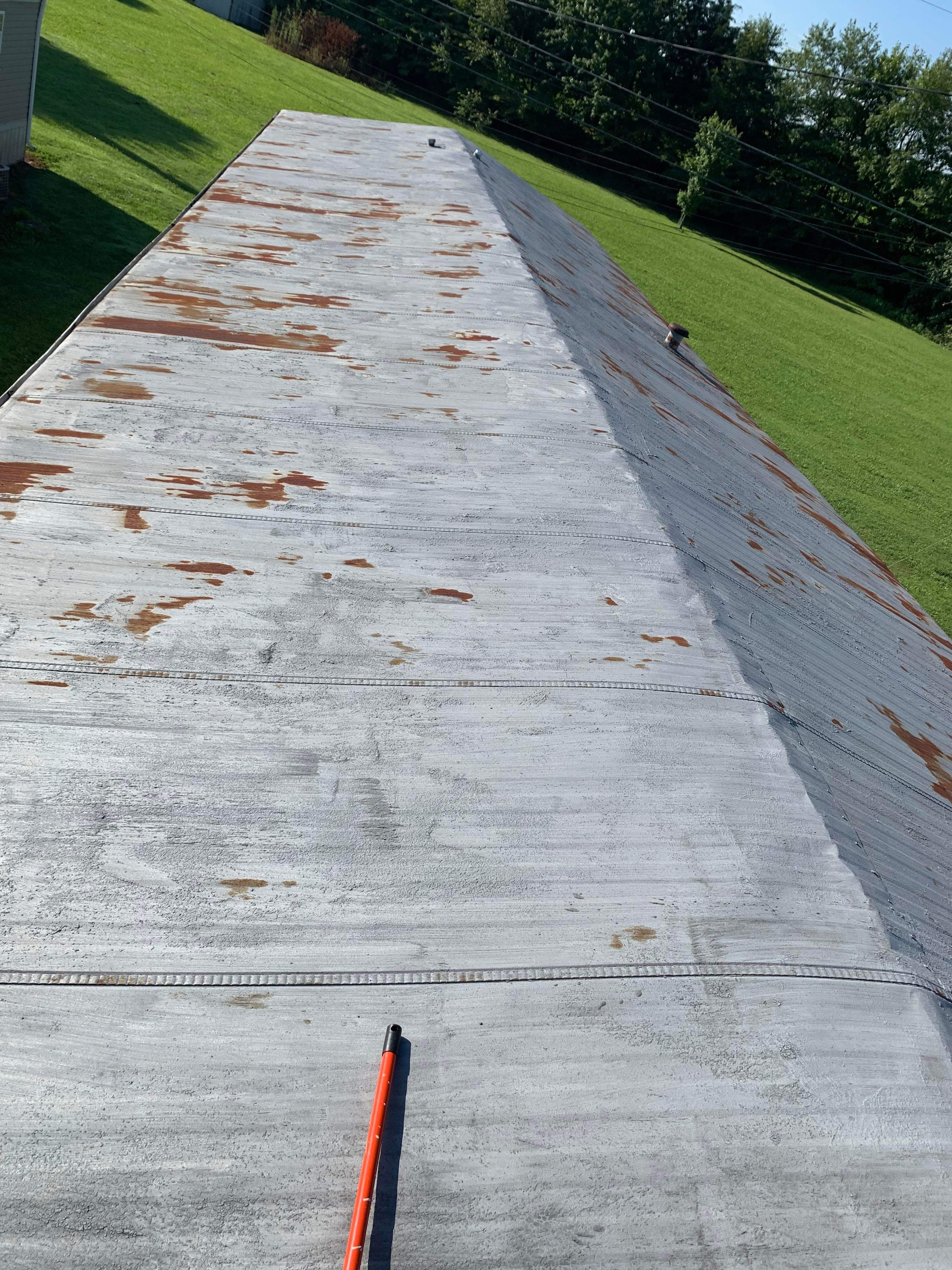 Mobile home metal roof before coating — heavily rust-stained and weathered silver metal panels with orange rust patches and a paint roller on the surface